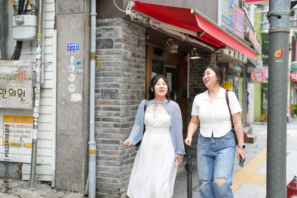 Fototapeta premium Two Korean women in their twenties in casual clothes walk along a boulevard sidewalk, talking and smiling as the city awakens under bright spring light, passing shops. Seoul, Jungu District, spring.