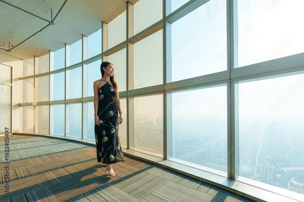Fototapeta premium Beautiful asian stylish woman in elegant dress standing in front of glass window inside modern highrise building with city skyline at Bangkok