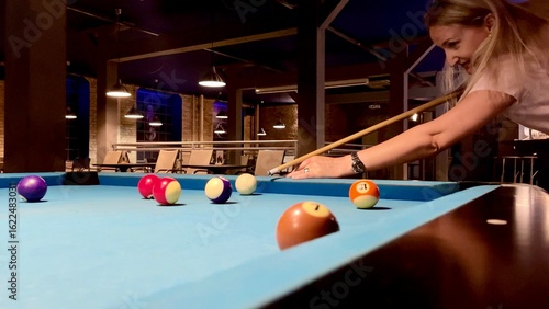 Middle-age blonde woman holding arm on billiard table, playing snooker game or preparing aiming to shoot pool balls. sport game snooker billiards