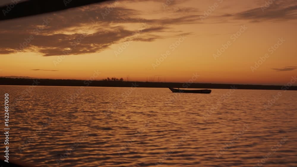 Lonely boat on water's surface with slight ripple at sunset. Coast is covered with grass and trees under soft golden sky