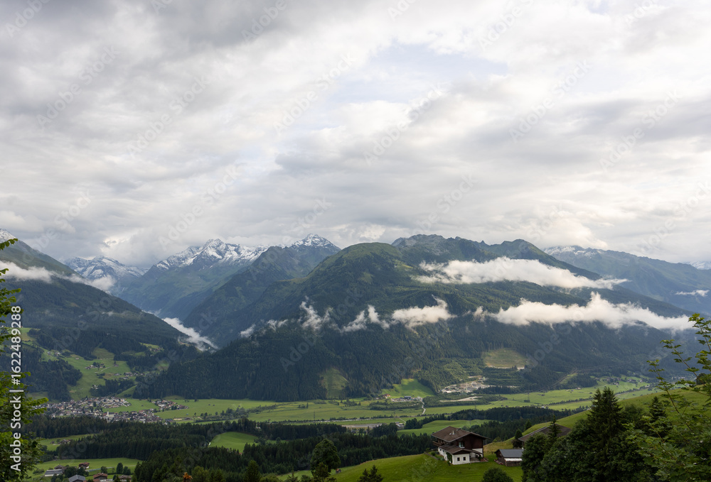 Fototapeta premium Breathtaking alpine valley with low clouds, green slopes, forested mountains, and distant snowy peaks in summer