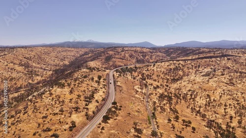 Aerial view of a road winding through arid land towards Lassen peak, a stark contrast to the distant snow-capped mountains under a clear sky, Red Bluff, California, United States.