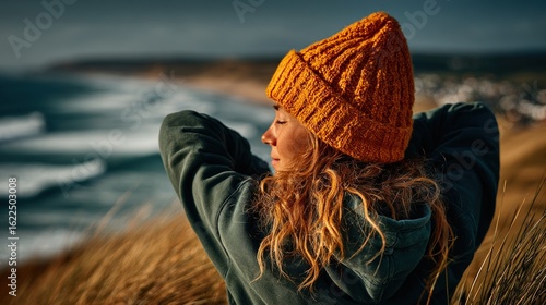 Woman in a yellow beanie and green hoodie looking out at the ocean, hands behind head, enjoying the beach scene.