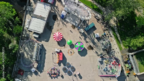 Aerial top-down drone video zooming out from a vibrant amusement park at City Park Ice Rink & Boating, Budapest. Morning light highlights colorful rides and greenery in this dynamic urban scene.