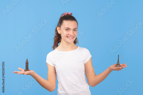 Smiling teen girl holding Eiffel Tower and Big Ben miniatures. Travel and culture comparison concept