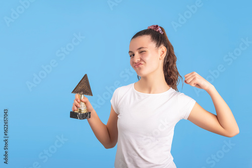 Proud teen girl flexing her arm and holding a trophy. Concept of strength, victory, empowerment and goal achieved