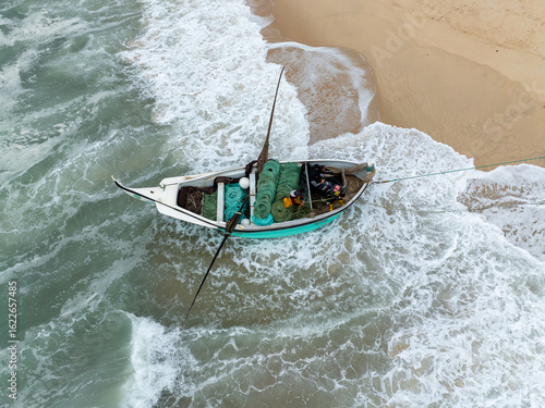 Aerial view of a small boat with fishing nets stranded on the shore, caught between the sand and the turbulent sea, Praia de Mira, Coimbra, Portugal.