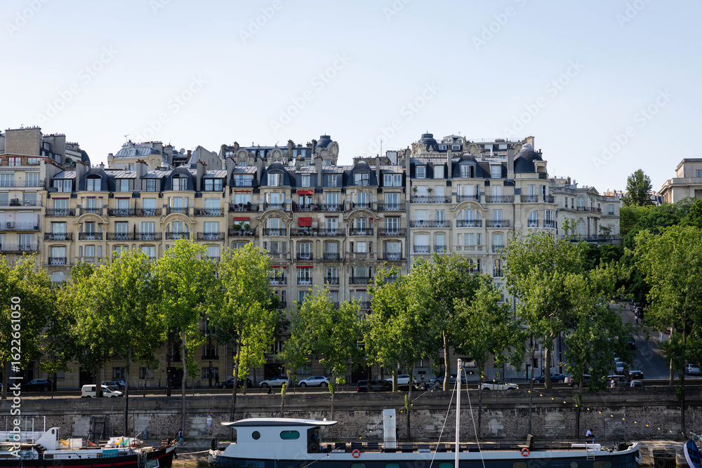 Naklejka premium Parisian apartment buildings, classic Haussmann architecture, tree-lined street, summer day, tranquil mood, Seine River view, boats moored, urban landscape photography.