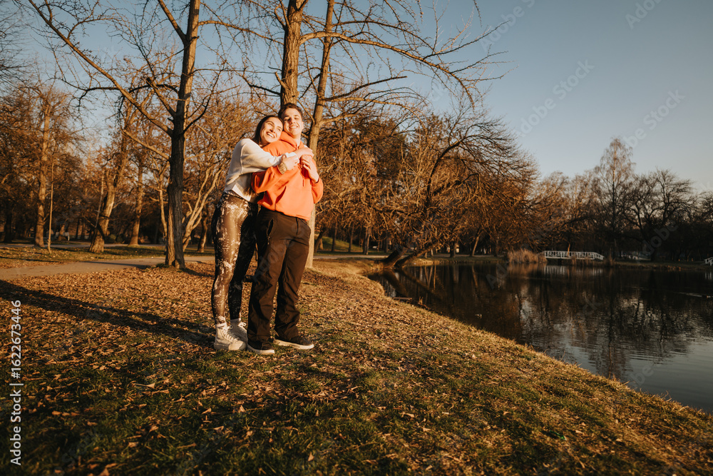Fototapeta premium A couple enjoying a cozy moment together under golden sunlight by a serene park lake, surrounded by trees capturing the essence of autumn.