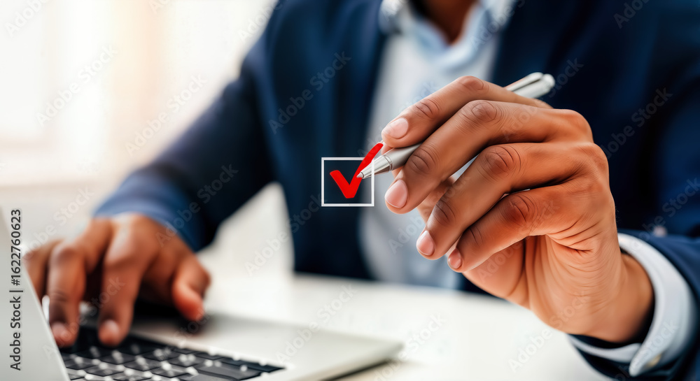 Obraz premium A businessmans hand holding a pen, marking a checkmark in a box on a document while working on a laptop in an office setting, symbolizing task completion and decisionmaking