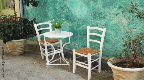 Wallpaper Mural Charming outdoor café corner in Aegina, Greece with two white chairs, round table and blooming plant. Straw seats and turquoise wall add calm, welcoming feel. No people, summer light Torontodigital.ca