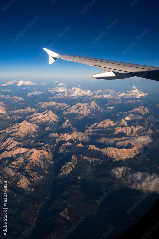 Obraz premium Airplane wing view over mountain range with blue sky and clouds, aerial travel landscape from plane window.