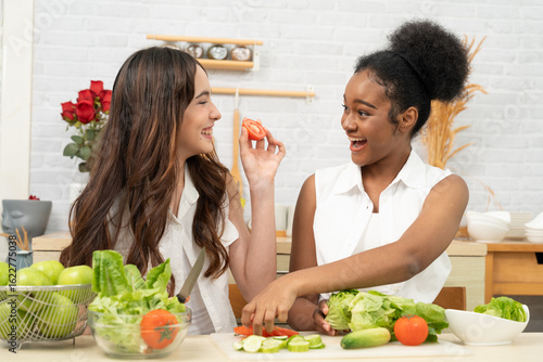 Black and white teenage friends happily and joyfully together to making a vegetable salad in a white kitchen. They enjoy healthy food.