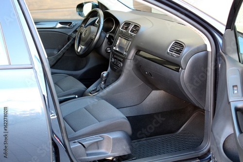 Passenger side view of a modern car interior with black fabric seats, central console with multimedia system, manual gear lever, soft plastic panels, and patterned rubber floor mats.