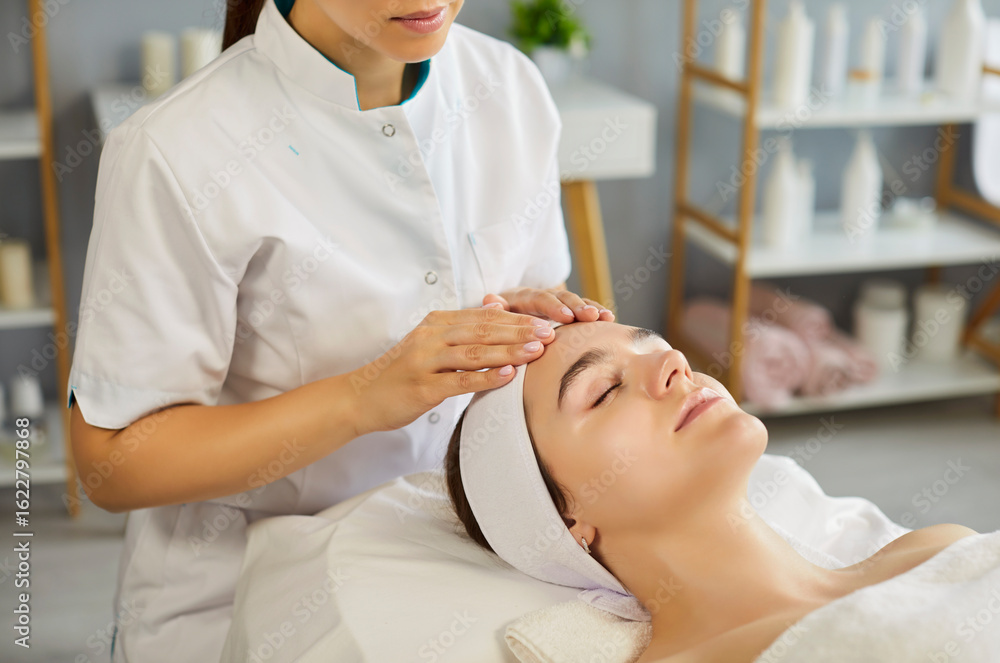 Obraz premium Professional female cosmetologist gently massages forehead of relaxed woman lying on a spa bed with closed eyes and headband. Clean clinic interior with skincare products in background.