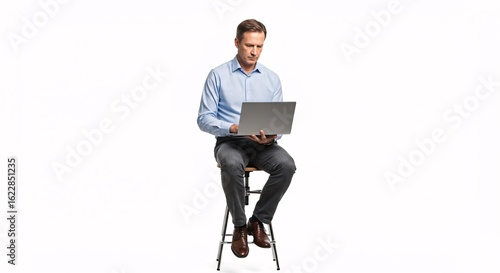 Focused middle-aged man sitting on a stool using a laptop. Wearing business casual attire with a blue shirt and dark pants, isolated on a white background.