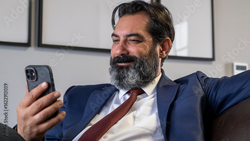 A middle-aged, dark-haired businessman in a suit is sitting on a leather couch in his office, happily engaged with his phone