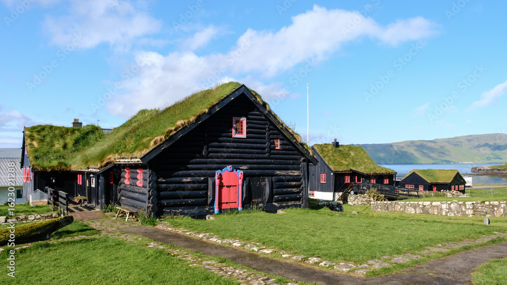 Fototapeta premium Quaint black wooden homes with grass roofs dot the lush green landscape of the Faroe Islands. Vibrant red doors and windows contrast beautifully with the surroundings. Kirkjubour village