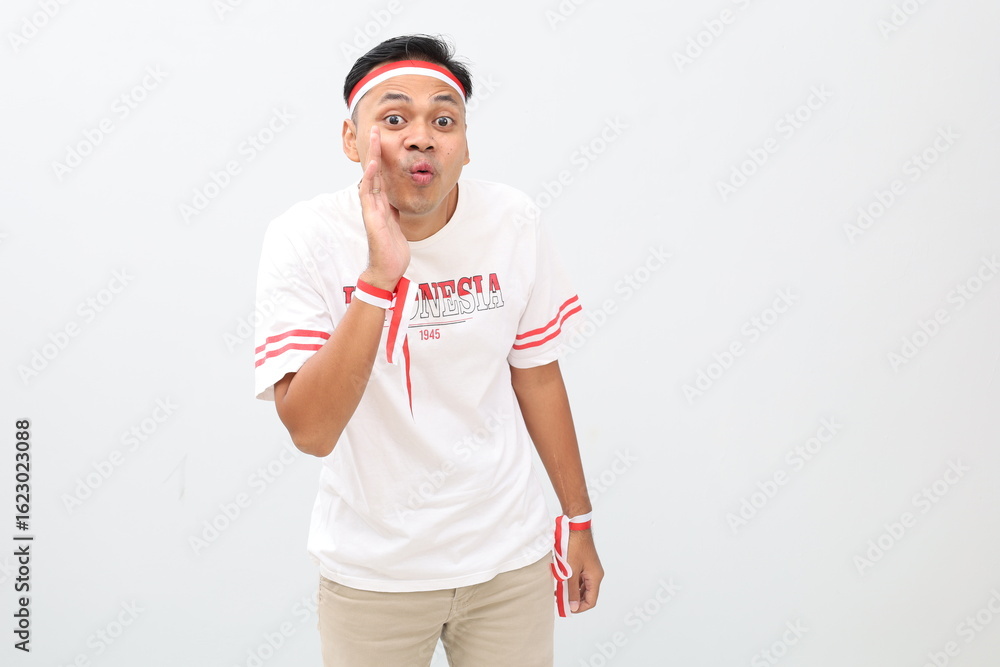 Naklejka premium Indonesian man with red and white ribbon celebrating Indonesian independence day, standing looking at camera with hand beside mouth, gesture of making announcement or whispering secret.