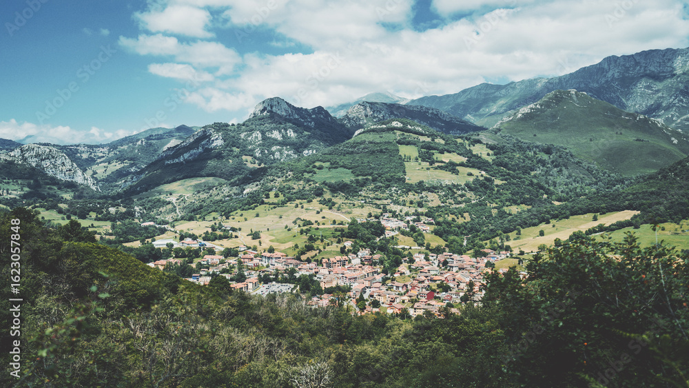 Obraz premium Picturesque view of Cabrales village nestled in lush green hills and dramatic limestone peaks of northern Spain, showcasing charming architecture and stunning mountainous landscape under a bright sky