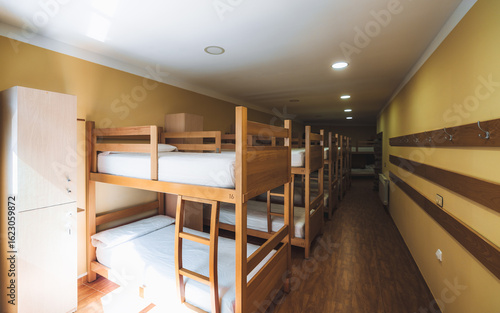 Clean and organized hostel dormitory room with multiple wooden bunk beds, individual lockers, wall hooks, and warm lighting, offering a functional shared sleeping space for travelers or pilgrims