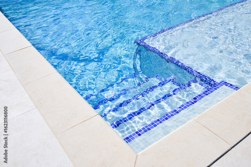 Obraz premium Stairs entrance to a private swimming pool with blue tiles and clear, calm water. The sunlight highlights the cleanliness, geometric lines, and refreshing atmosphere of the space.