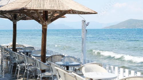 Wallpaper Mural Empty seaside café terrace with white tables and straw umbrellas by the water in Aegina, Greece. Clear sky, waves splashing near white railing, mountain silhouettes on horizon. Sunny day, no people. Torontodigital.ca
