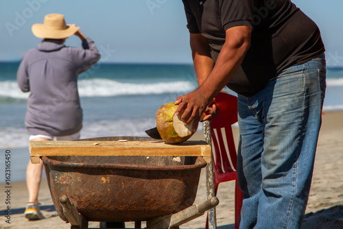 Close up of a man chopping a coconut on a beach with a machete. A perosn and the ocean are in the background