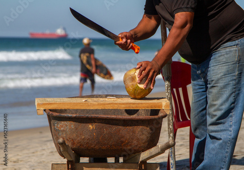 Close up of a man chopping a coconut on a beach with a machete. A perosn and a ship and the ocean are in the background