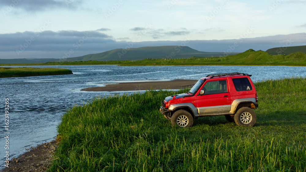 Red off-road vehicle parked on lush green grass by a serene riverbank, surrounded by rolling hills and a clear blue sky, showcasing adventure and exploration in nature Red off-road vehicle parked on lush green grass by a serene riverbank, surrounded by rolling hills and a clear blue sky, showcasing adventure and exploration in nature