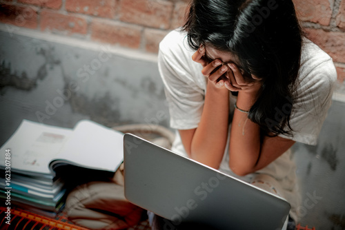 A young Asian teenage girl covering her face with both hands in front of a laptop while studying for high school final exam. Distress, frustration and mental health in education concept.