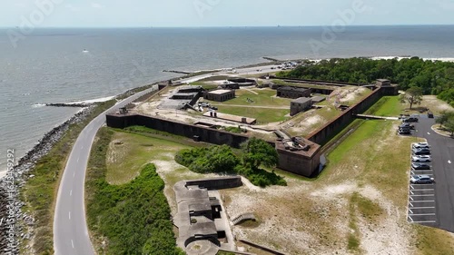 Aerial view of a historic fort surrounded by water, showcasing its large, star-shaped structure with multiple buildings and grassy areas. Nearby roads and parking areas are visible.