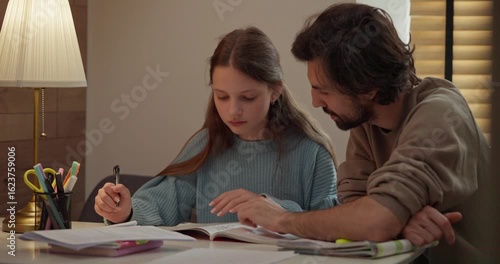 Teenage girl doing homework with her dad support sitting at table with desk lamp at home in the evening. Young father and daughter doing homework in the kitchen at home