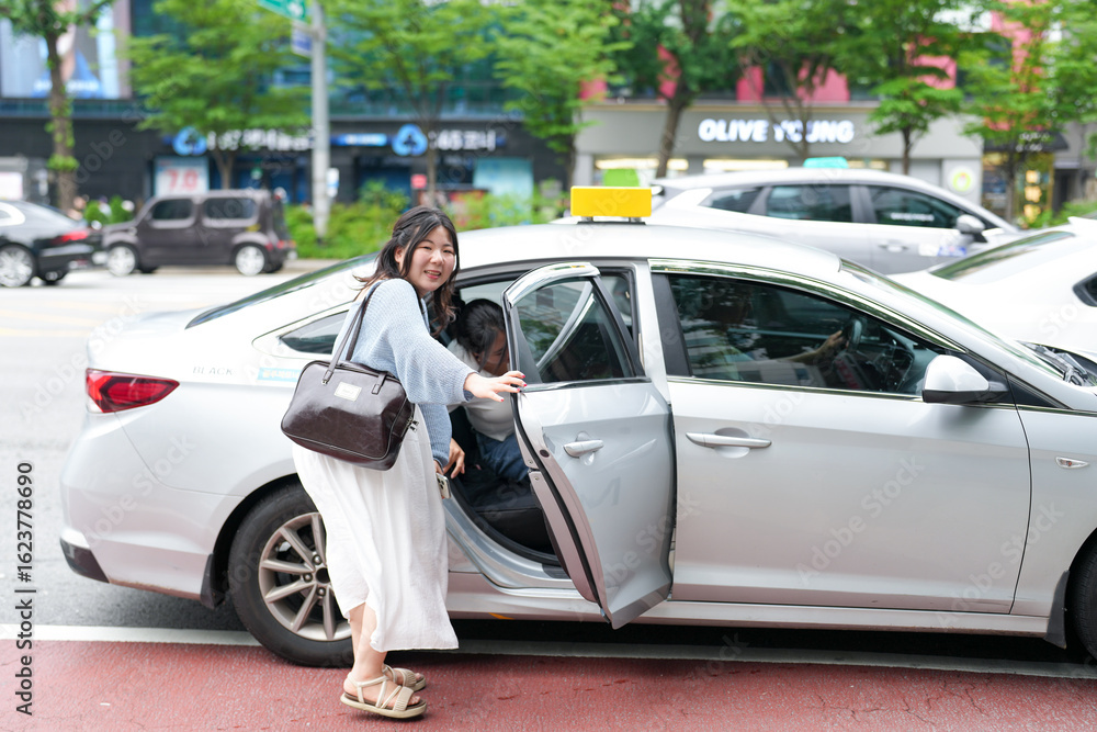 Fototapeta premium Two Korean women couple in their twenties in casual clothes step into a taxi on a busy boulevard, smiling and exchanging a glance as soft spring light fills the street. Seoul, Jung District, spring.