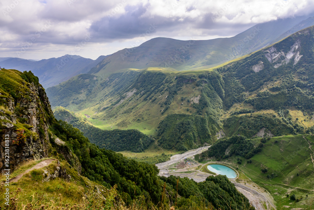 Fototapeta premium A scenic Caucasus vista showcases rugged peaks and lush valleys, with a small turquoise lake near Gudauri, Georgia.