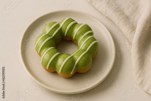 A round, bubble-shaped mochi doughnut glazed with a vibrant green tea coating and decorated with thin white icing stripes. Served on a neutral ceramic plate, minimalist setup highlights the doughnut