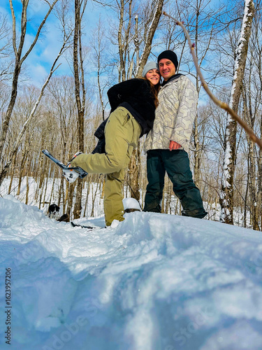 A couple takes a picture together snowshoeing in a snowy forest. The woman has her leg up with their dog in the background during the winter.