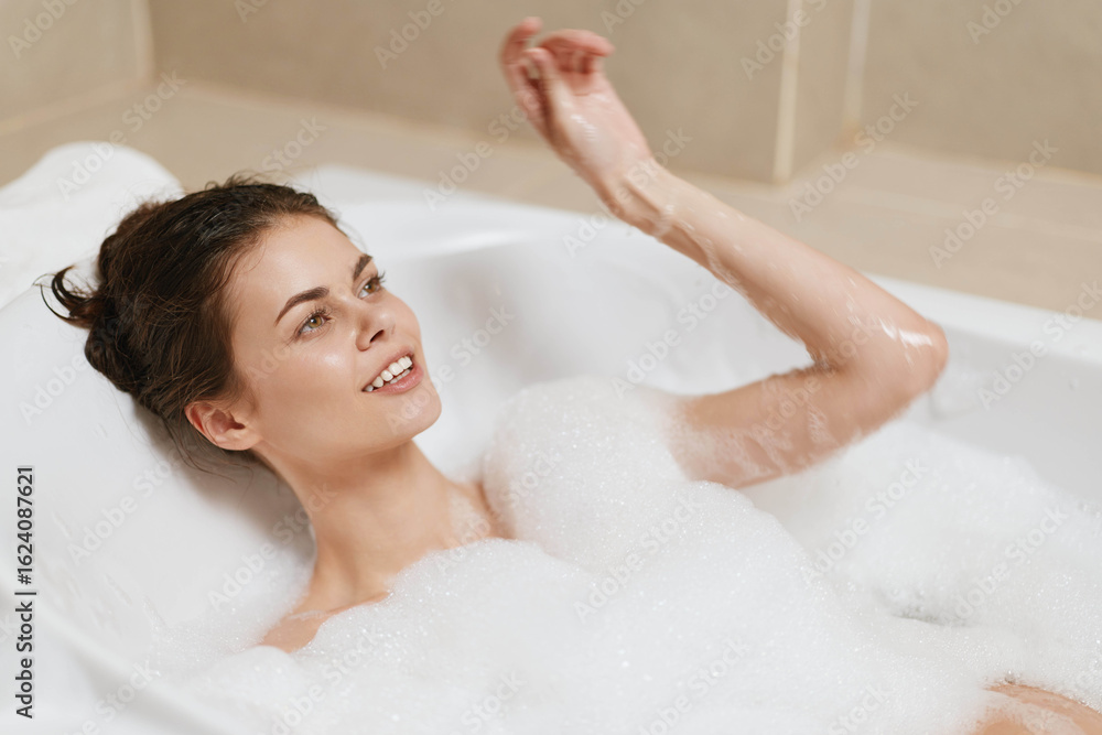 Obraz premium Relaxed young woman with a joyful smile enjoying a bubble bath, sitting in a modern tub with soft lighting and neutral-colored tiles in the background.