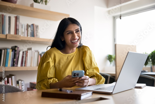 Foto Woman sit at desk hold smartphone, laughs on received sms, enjoy great news in m