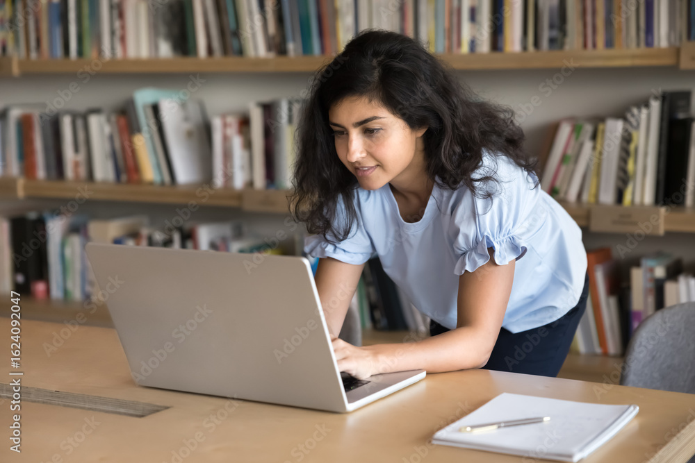 Indian student woman studying on laptop in college or high-school library, leaning at desk typing on device keyboard, prepare essay or assignment, working on online project, learn topic looks focused Indian student woman studying on laptop in college or high-school library, leaning at desk typing on device keyboard, prepare essay or assignment, working on online project, learn topic looks focused