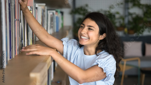 Billede på lærred Happy Indian woman enthusiastically reaching for book on shelf, reflects curiosi