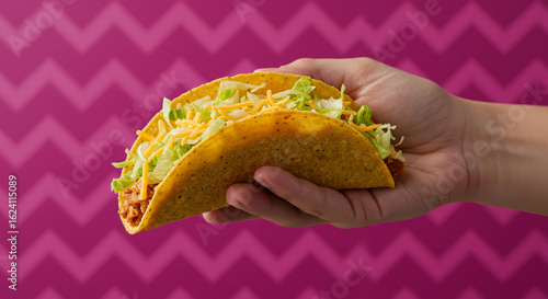 ''A hand holding a vibrant, appetizing hard-shell taco filled with shredded lettuce, cheese, and seasoned meat against a bold p...