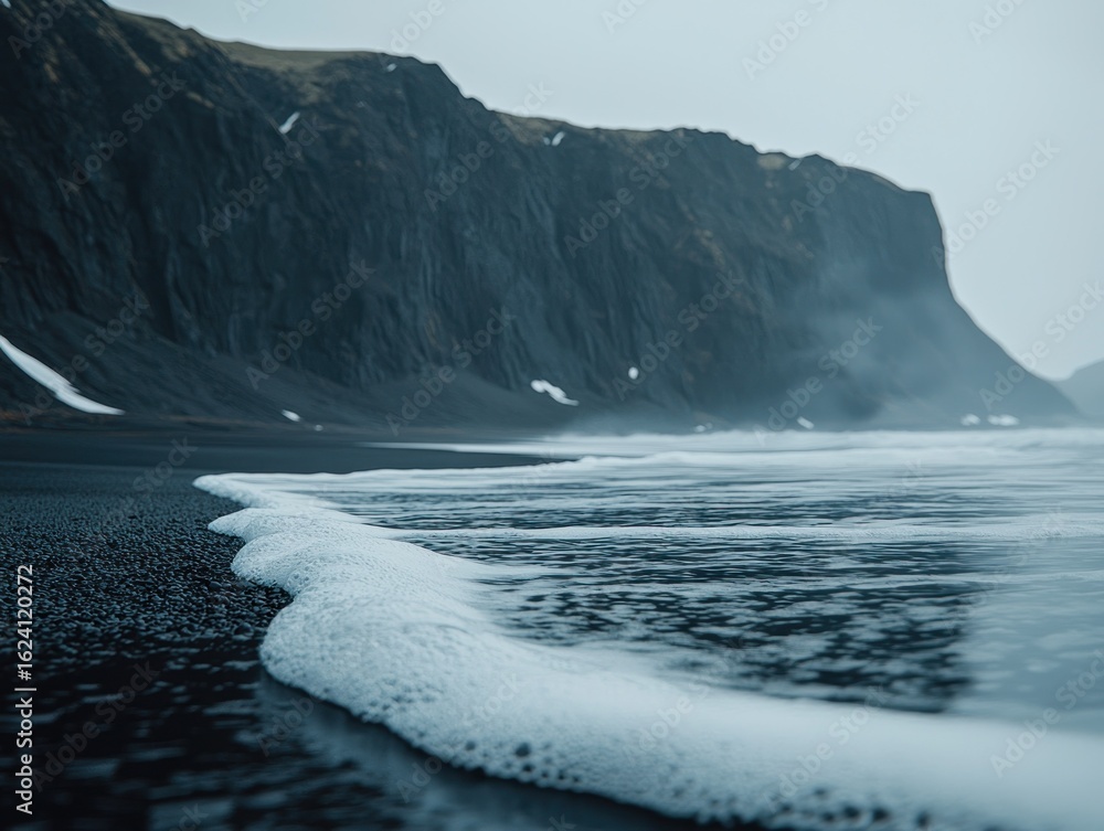 Naklejka premium Foamy waves lapping at a black sand beach, dramatic cliffs loom in the background