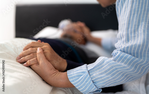 scene of bedridden patient, focus on hand of wife holding hands illness elderly hopeless senior man. home caregiver nurse visit retirement patient for health check up recovery process medical service