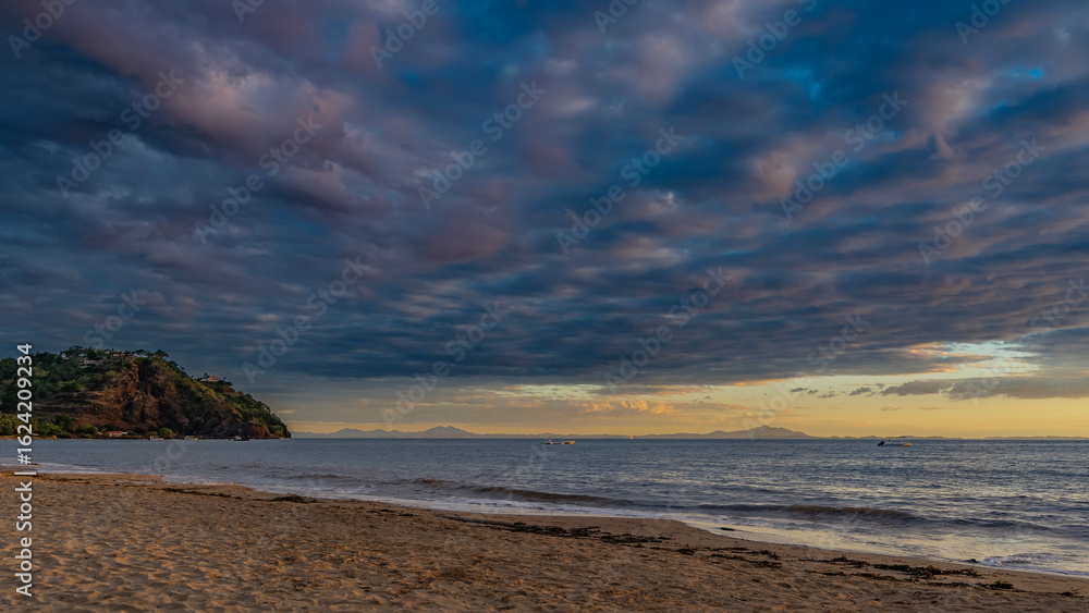 Fototapeta premium Sunset on a tropical island. Ocean waves are foaming on a sandy beach. Blue clouds are highlighted in pink. The sky is orange on the horizon. A hill in the distance. Madagascar. Nosy Be 