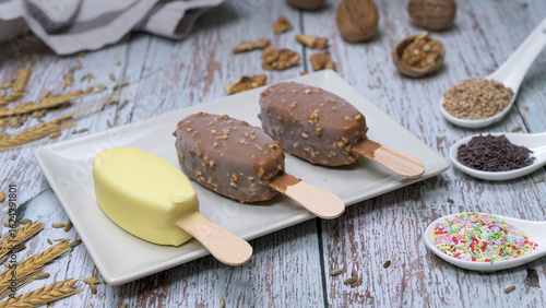 Delicious scene with three ice cream bars, including chocolate and vanilla flavors, served on a white plate with toppings, seeds, nuts, and colorful sprinkles. Rustic wooden background 