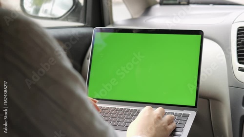 Person is using a laptop in a car. The laptop screen is green. The person is typing on the keyboard