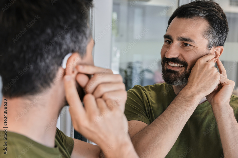 Fototapeta premium Man with hearing problems tests out his new behind-the-ear hearing aid, smiling as he observes himself in home mirror. Audiology