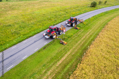 Drone view of two tractors with a mechanical mowers mowing grass on the side of the asphalt road
