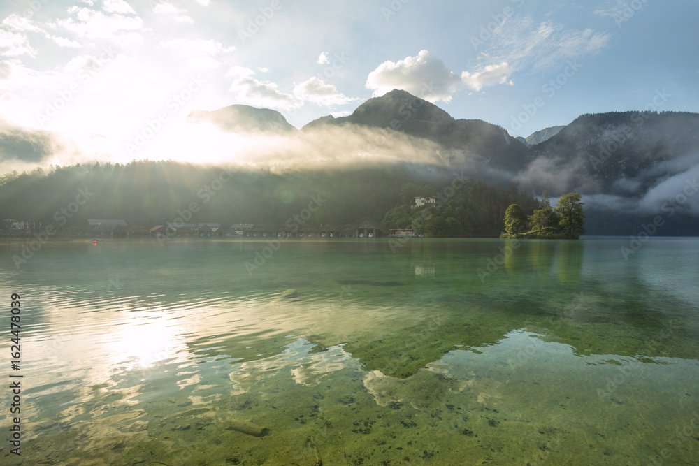 Naklejka premium Mystischer Morgen am Königssee in Schönau mit Bootshäusern und Christlieger. Sonnenaufgang und schöne Nebelschwaden über dem Königssee im Kessel.